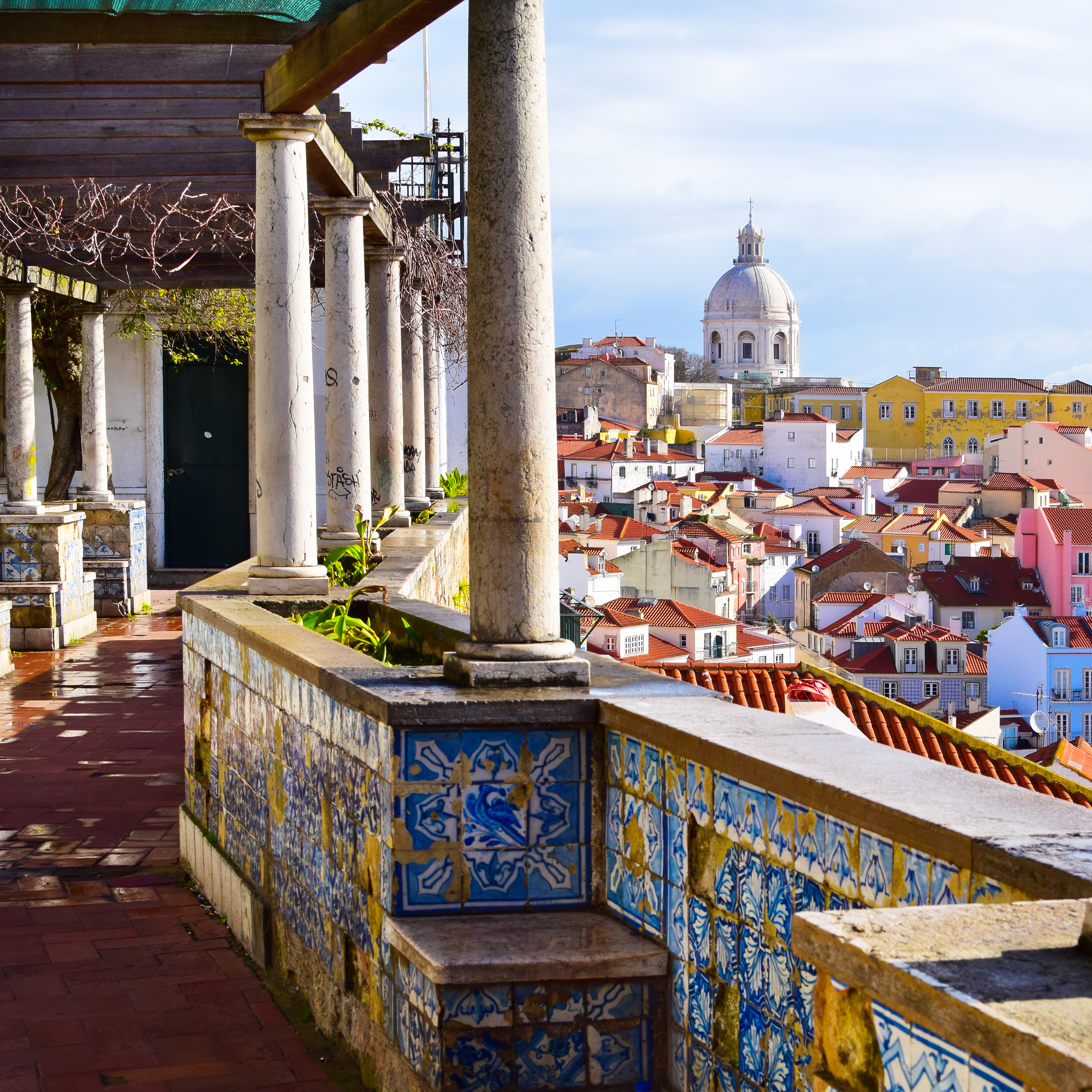 Lisbon cityscape from a hilltop miradouro — terracotta rooftops and the Tagus estuary stretching to the horizon